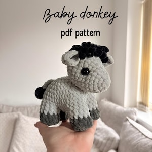 May include: A crocheted grey and black baby donkey plush toy. The toy has a black mane and tail. The text "Baby donkey pdf pattern" is written above the toy.