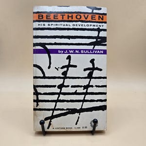 May include: A vintage book titled "BEETHOVEN HIS SPIRITUAL DEVELOPMENT" by J.W.N. Sullivan. The book cover is white with black abstract musical notes and the title in orange and black. A purple banner displays the author's name.
