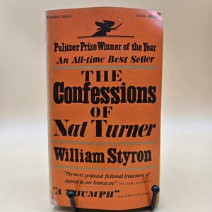 May include: An orange paperback book titled "The Confessions of Nat Turner" by William Styron. The cover features black text and a graphic of a person with a sword. The book is a Pulitzer Prize Winner and an All-time Best Seller.