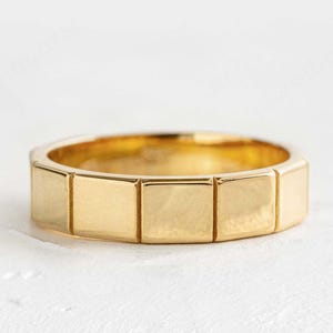 Puede incluir: Un anillo de oro con un diseño geométrico. El anillo presenta una serie de segmentos rectangulares alrededor de la banda. El anillo está hecho de un material de oro amarillo brillante y está sobre un fondo blanco. Joyería.