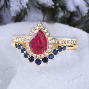 Op de afbeelding: Een driedelige gouden ringenset. De verlovingsring heeft een peervormige, dieprode robijn omringd door kleine diamanten. De trouwring heeft kleine diamanten en de derde ring heeft blauwe saffieren.