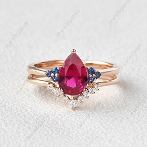 Op de afbeelding: Een verlovingsring met een peervormige robijn, omringd door een halo van kleinere diamanten en twee blauwe saffieren aan elke kant. De ring is gemaakt van roségoud.