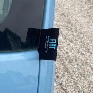 May include: A black Fiat tag with the Fiat logo in light blue. The tag is attached to a light blue car door. The background is a light brown, textured surface.