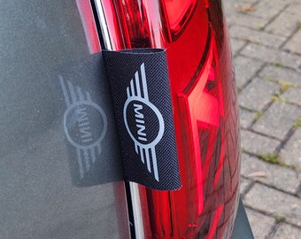 May include: A black fabric patch with the word "MINI" in a white circle and a silver wing design. The patch is attached to a red taillight on a car.