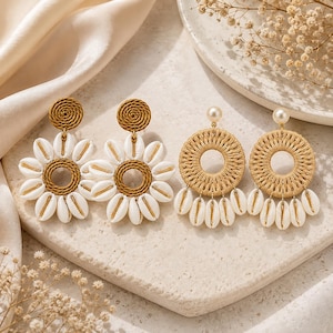 Peut inclure: Deux paires de boucles d'oreilles. La première paire présente un clou circulaire tressé marron avec un pendentif en forme de fleur composé de coquillages cauris blancs autour d'un centre tressé marron. La seconde a une perle blanche.