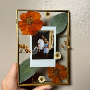 以下が含まれることがあります： 金色のフレームに押し花のデザインが施された写真。写真には、部屋でイルミネーションをバックにポーズをとるカップルが写っている。花はオレンジ、白、緑の色合いで構成されている。