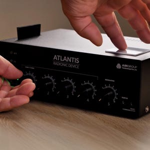 Puede incluir: Un "ATLANTIS Radionic Device" negro con múltiples diales y una pequeña placa rectangular en la parte superior. El dispositivo tiene el logotipo "AIM Group Technology for Life". Las manos ajustan los diales y tocan la placa.