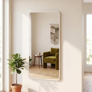 May include: A tall, rectangular wall mirror with a thin, white frame and rounded corners. The reflective surface shows a room interior with a green armchair, a small wooden side table, and a wall hanging, providing a clear, undistorted view.