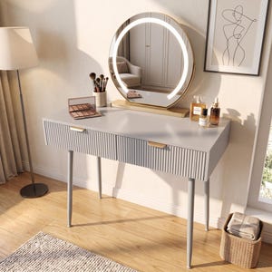 May include: A grey vanity table with two drawers featuring vertical ribbed fronts and gold-toned handles. A round, gold-framed mirror with an integrated LED light ring sits on top, accompanied by makeup brushes, a palette, and perfume bottles.