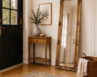 May include: A tall, rectangular floor mirror with an ornate, metallic gold frame featuring intricate carvings. The mirror stands upright against a white wall, reflecting a doorway and interior space, positioned next to a woven basket with a white cloth.