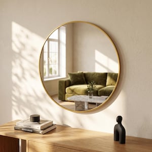 May include: A circular wall mirror featuring a thin, metallic gold frame. The reflective surface displays a room interior with a green sofa, a marble coffee table, and a window. The mirror is mounted on a plain wall above a wooden surface.
