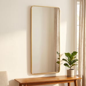 May include: A rectangular wall mirror featuring a thin, metallic gold frame with rounded corners. The mirror has a reflective glass surface that shows a partial reflection of a green potted plant and a window with light-coloured curtains.
