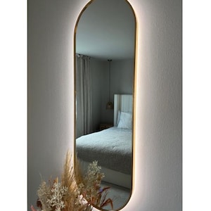 May include: An oval mirror with a gold frame and integrated lighting, mounted on a white wall. The mirror reflects a bedroom scene. The frame has a warm, golden colour, and the lighting creates a soft glow around the edge. A bouquet of dried flowers is visible below.
