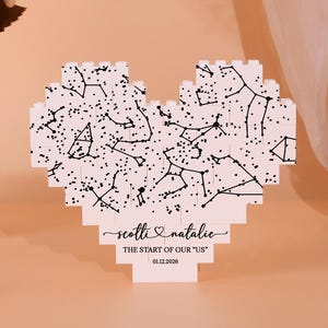 Op de afbeelding: Een wit, hartvormig houten puzzel met een sterrenbeeldontwerp met zwarte lijnen en stippen. De puzzel draagt de namen "Scottie & Natalie" en de tekst "THE START OF OUR US" met de datum 01.12.2020. Gepresenteerd op een perzik-gekleurd oppervlak.