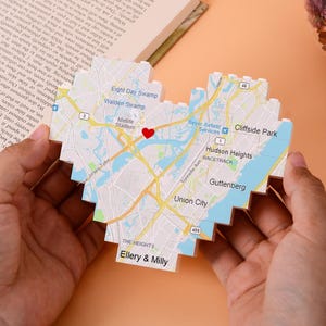 Puede incluir: Un mapa en forma de corazón de una ciudad, sostenido en las manos. El mapa presenta carreteras, puntos de referencia y el texto "Ellery & Milly" en la parte inferior. Un corazón rojo señala una ubicación. El mapa está hecho de pequeños bloques pixelados.