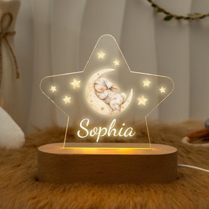 Op de afbeelding: Een houten nachtlampje in de vorm van een ster met een halve maan en een slapende baby erin. De ster wordt verlicht met warmwitte LED-lampjes en de naam "Sophia" staat onder de maan.