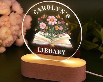 May include: A personalised night light with a wooden base. The clear, circular light features the text "CAROLYN'S LIBRARY" and a floral design above an open book. The light emits a warm glow.
