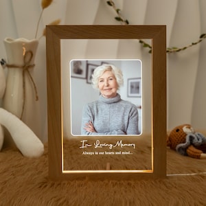 Puede incluir: Marco de fotos LED de madera con una foto de una mujer. El marco tiene el texto "In Loving Memory" y "Always in our hearts and mind..." en una fuente de escritura. El marco está iluminado con un brillo cálido.