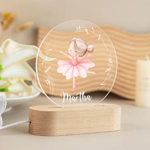 May include: A personalised night light featuring a ballerina in a pink tutu, with musical notes around the edge. The name "Martha" is written below the ballerina. The light sits on a wooden base.