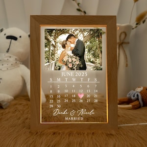 Op de afbeelding: Houten omlijste verlichte kalender met een trouwfoto. De kalender toont juni 2025, met de namen "Drake & Nicole" en het woord "MARRIED" eronder. De lijst is gemaakt van lichtbruin hout.