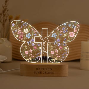 May include: A butterfly-shaped night light with floral wings and a cross design. The cross features the name "Sarah" and is centered on the wings. The wooden base displays the text "BAPTIZED JUNE 28, 2025".
