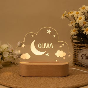 May include: A white acrylic night light shaped like a cloud with a crescent moon, stars, and the name "Olivia" inscribed. The light sits on a wooden base.