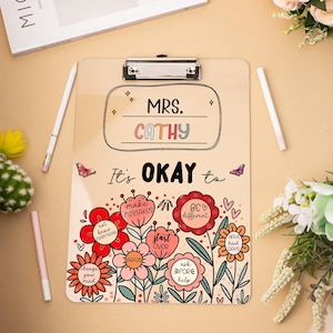 Op de afbeelding: Een klembord met een metalen clip, met de naam "CATHY" en de zin "It's OKAY to" met bloemenillustraties en inspirerende boodschappen. Het ontwerp bevat vlinders en verschillende bloemvormen met positieve affirmaties.