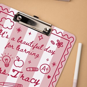 May include: A pink and white striped clipboard with the text "It's a beautiful day for learning" and "Mrs. Tracy" in a handwritten font. The clipboard has a silver metal clip and decorative elements like stars, an apple, and a lightbulb.