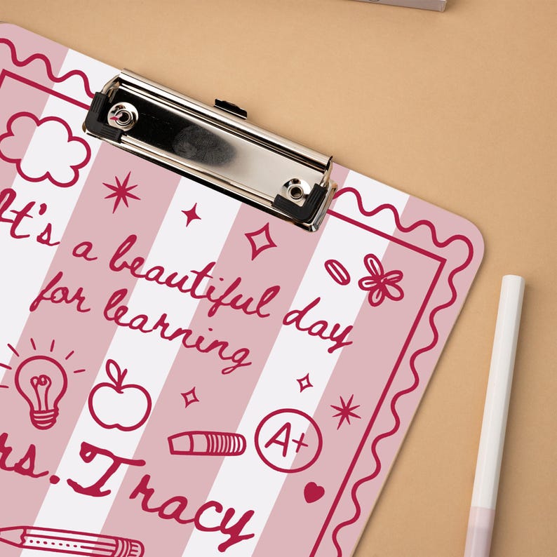 May include: A pink and white striped clipboard with the text "It's a beautiful day for learning" and "Mrs. Tracy" in a handwritten font. The clipboard has a silver metal clip and decorative elements like stars, an apple, and a lightbulb.
