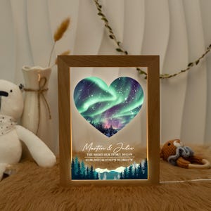 May include: A wooden framed night light with a heart-shaped design featuring a green and purple aurora borealis scene. The text reads "Martin & Julie, The Night Our Story Began, 03.08.2025 44.97135&deg;N 93.28611"W". The bottom of the light has a forest scene.