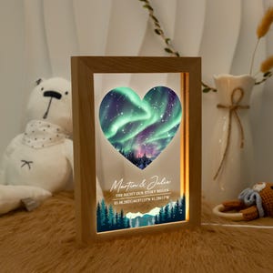 May include: Wooden framed night light with a heart-shaped design featuring a green and purple aurora borealis scene. The text reads "Martin & Julie", "THE NIGHT OUR STORY BEGAN", and the date and coordinates. The light is on.
