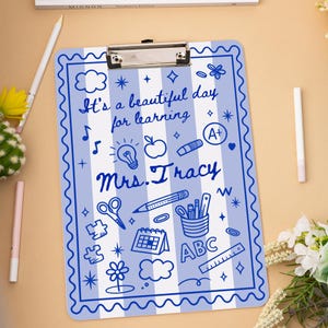 May include: A blue and white striped clipboard with a decorative border. The clipboard features the text "It's a beautiful day for learning" and "Mrs. Tracy" along with various school-themed doodles.