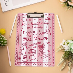 May include: A pink and white striped clipboard with the text "It's a beautiful day for learning Mrs. Tracy" and various school-themed doodles. Includes a metal clip and is surrounded by pens and flowers.