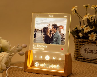 Pode incluir: Um tocador de música de madeira com um painel de acrílico transparente que exibe uma foto de um casal e o título da música "La Bachata" de Manuel Turizo. O tocador tem um ícone de coração, botões de reprodução e pausa e uma barra de progresso.