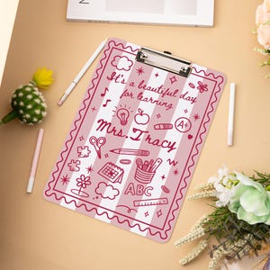 May include: Pink and white striped clipboard with the text "It's a beautiful day for learning Mrs. Tracy" and various school-themed doodles. The clipboard has a silver metal clip at the top.