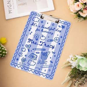 May include: A blue and white striped clipboard with the text "It's a beautiful day for learning Mrs. Tracy" and various school-themed illustrations. The clipboard has a silver metal clip at the top.