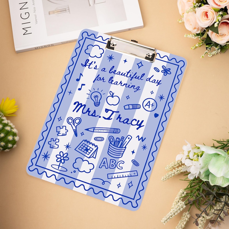May include: A blue and white striped clipboard with the text "It's a beautiful day for learning Mrs. Tracy" and various school-themed illustrations. The clipboard has a silver metal clip at the top.