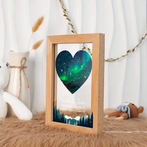 May include: A wooden framed print featuring a heart-shaped image of the night sky with green and white aurora borealis. The print includes the text "Martin & Julie" and the date and coordinates of a special event. The frame is made of light-colored wood.