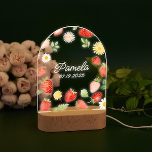 Peut inclure: Une veilleuse personnalisée avec une base en bois et un dessus en acrylique en forme d'arche. Le dessus est décoré de fraises, de marguerites et du nom "Pamela" avec la date "07.19.2025". La lumière émet une douce lueur.