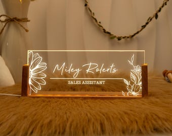 May include: A wooden desk sign with a clear acrylic panel featuring a floral design and the text "Miley Roberts, Sales Assistant". The sign is illuminated with warm white LED lights.