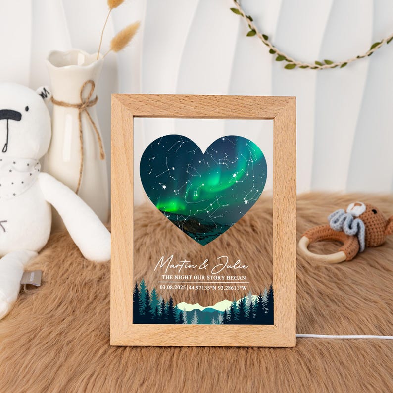 May include: A framed print featuring a heart-shaped image of the aurora borealis and constellations. The print includes the names "Martin & Julie" and the text "THE NIGHT OUR STORY BEGAN" with a date and coordinates. The frame is made of light-colored wood.