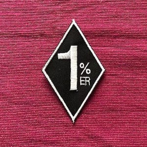 Könnte beinhalten: Schwarz-weißer, gestickter Aufnäher in Diamantenform. Der Aufnäher zeigt die Zahl "1" und den Text "% ER" in Weiß. Der Aufnäher befindet sich auf einem strukturierten, tiefrosa Stoffhintergrund.