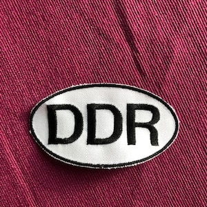 Könnte beinhalten: Weißer ovaler Patch mit schwarzem gesticktem Text "DDR" auf einem burgunderfarbenen Stoffhintergrund.