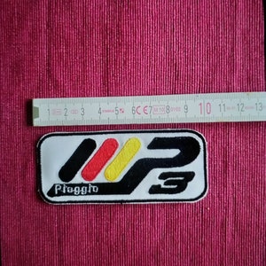 Könnte beinhalten: Weißer gestickter Aufnäher mit einem stilisierten "P" und "3" in Schwarzweiß mit einem roten, gelben und schwarzen Streifenmuster. Der Text "Piaggio" befindet sich unter dem Design.