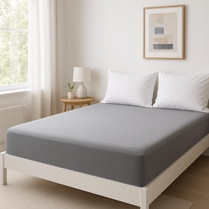 Könnte beinhalten: Ein graues Spannbettlaken auf einem Bett mit weißen Kissenbezügen. Das Bett hat einen Holzrahmen und steht auf einem hellgrauen Teppich.
