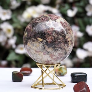 Puede incluir: Una esfera pulida de jaspe rosa, negro y blanco sobre un soporte de metal dorado. La esfera tiene un diámetro de aproximadamente 5 cm.