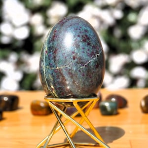 Könnte beinhalten: Ein grünes und rotes Edelsteinegg auf einem goldenen Metallständer. Das Ei ist glatt und poliert und hat eine marmorierte Optik.