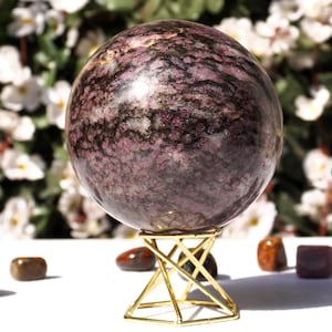 Puede incluir: Una gran esfera pulida de color púrpura y negro sobre un soporte de metal dorado. La esfera está hecha de piedra natural y tiene un patrón de remolino.