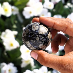 Könnte beinhalten: Eine schwarze und silberne Kugel mit einem marmorierten Muster wird in einer Hand gehalten. Die Kugel ist etwa so groß wie ein Golfball.