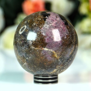 Könnte beinhalten: Eine polierte Edelstein-Kugel mit braunen, violetten und goldenen Farbtönen. Die Kugel steht auf einem schwarz-silbernen Sockel. Der Hintergrund ist unscharf, mit weißen und orangefarbenen Blumen.